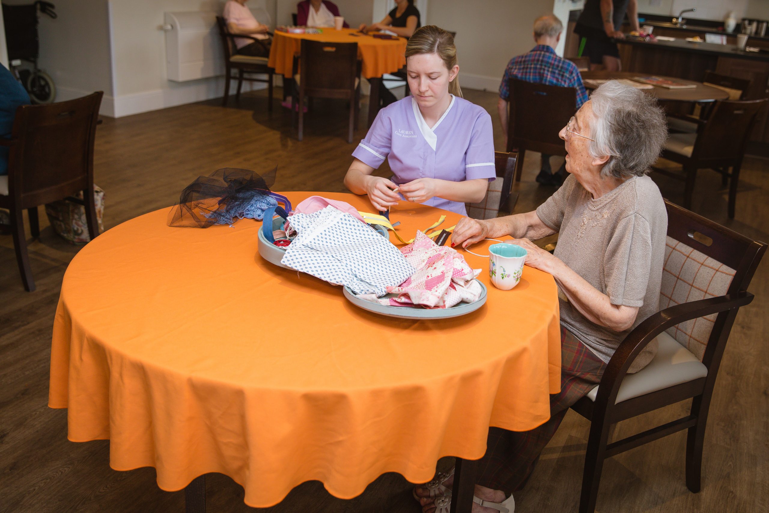 A resident enjoying some arts and crafts on a orange covered table with a female carer wearing purple uniform.