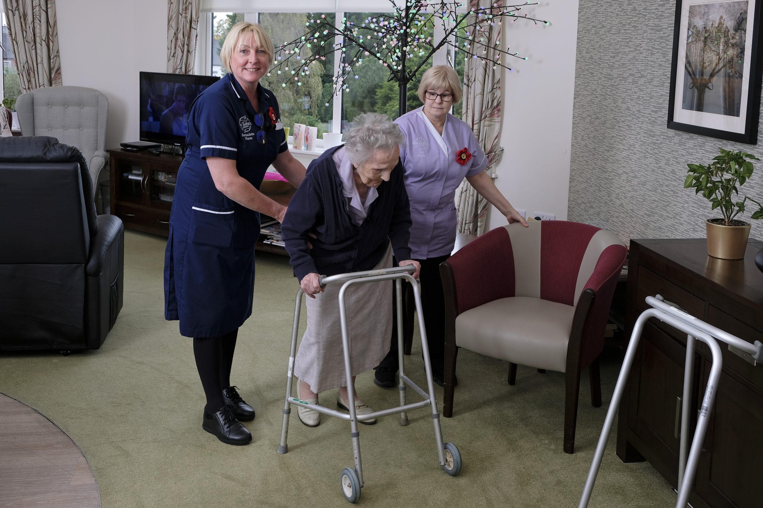 A female resident walking with a frame being helped by a care assistant and a nurse.