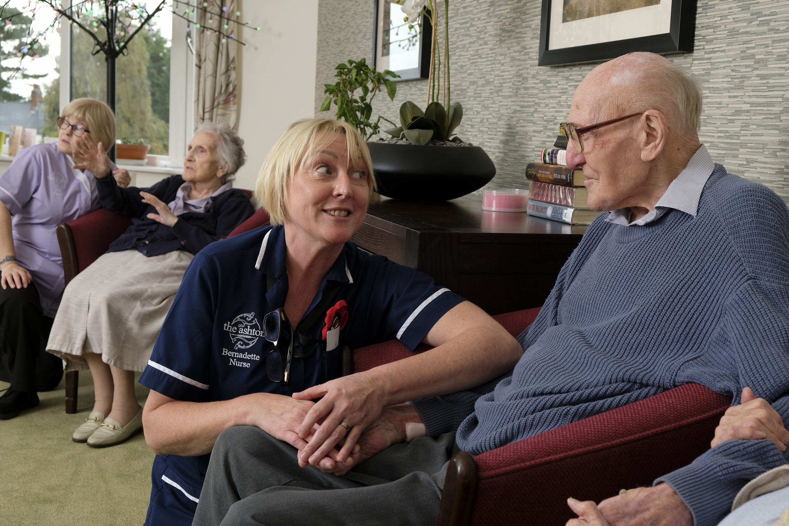 A nurse speaking to a male resident and a care assistant talking to a female resident in the background.