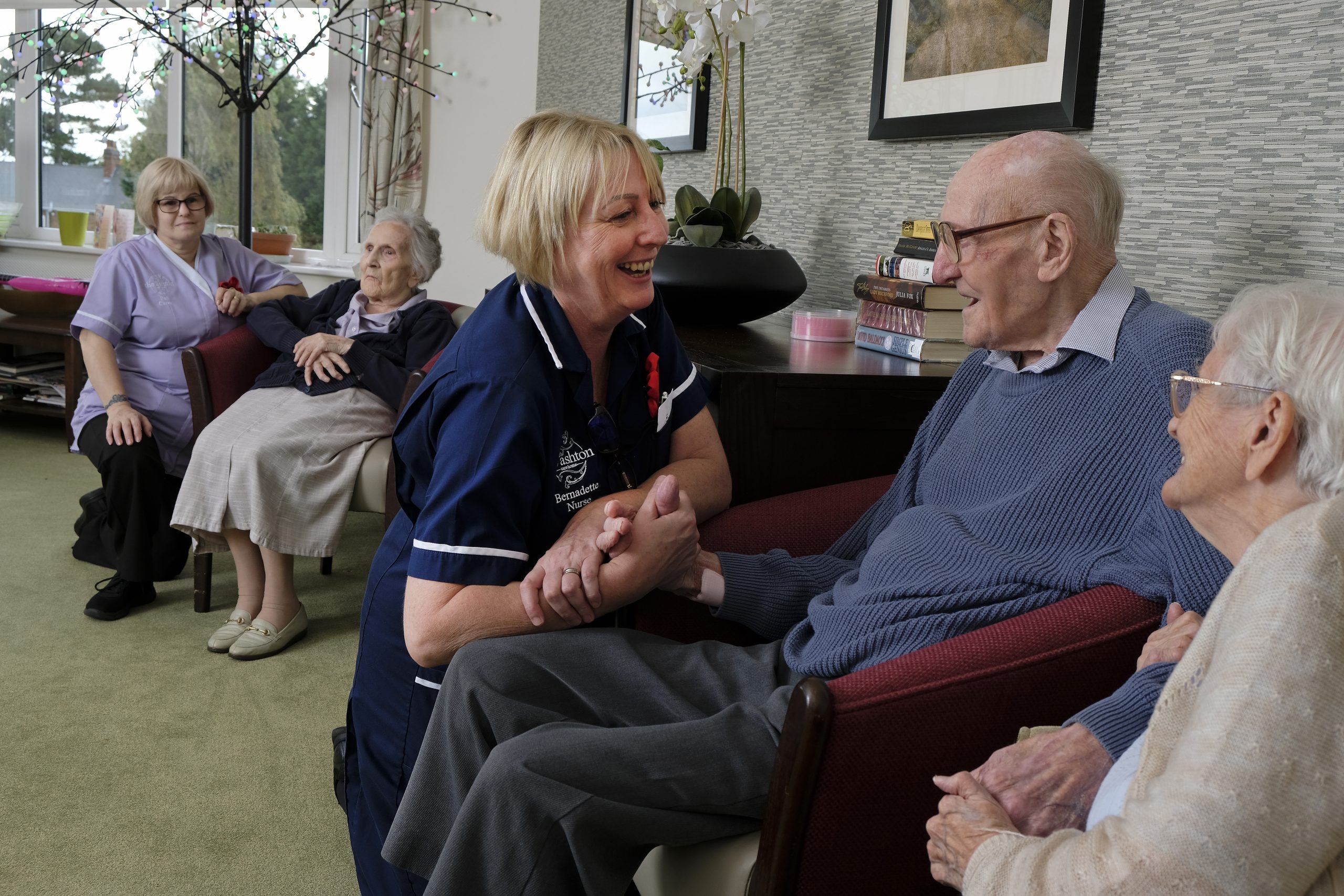 A nurse smiling with our residents who are holding hands. In the background, a carer talking to a female resident.