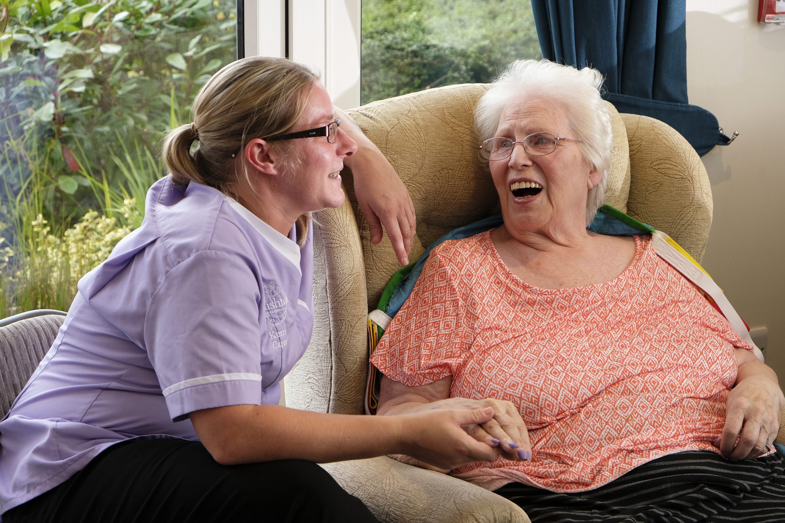 A caregiver wearing purple laughing with a female resident sat in a chair.