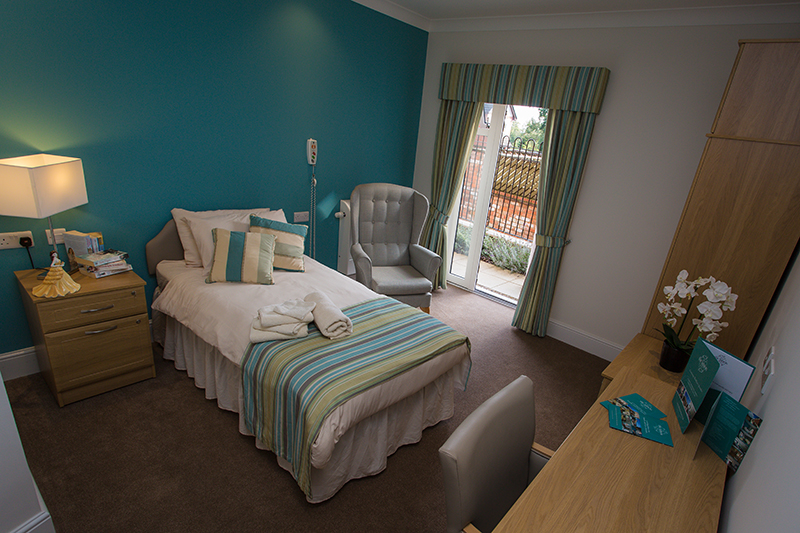 Bed with striped pillow and throw with matching curtains. Grey armchair. Grey carpet. Blue feature wall. Bedside table with lamp.