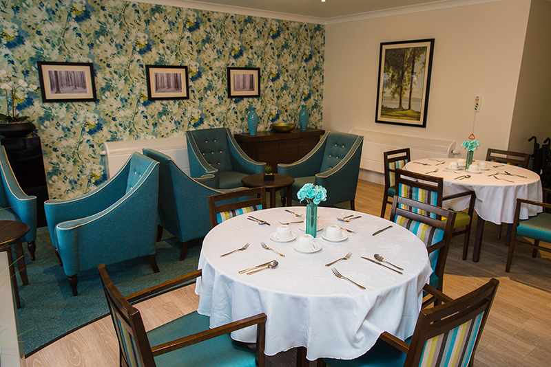 Dining room with two dressed tables. Two seating areas with blue chairs and dark brown coffee tables. Floral blue wallpaper.