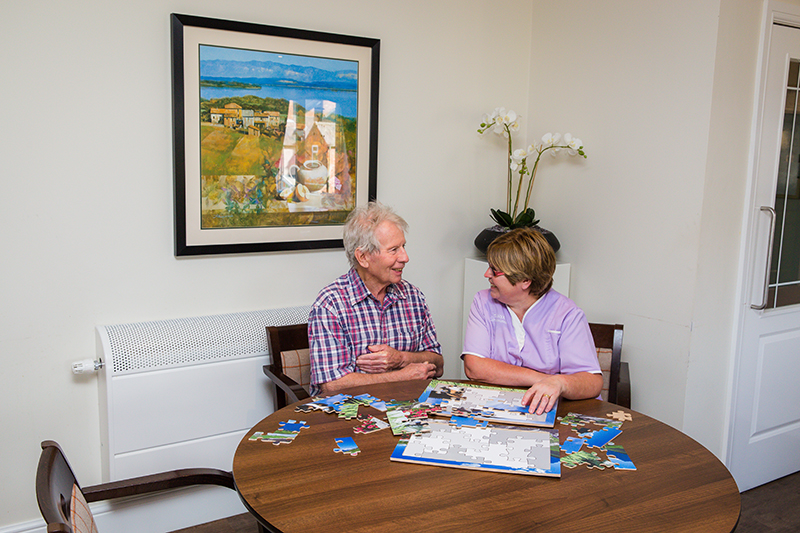 A male resident and a female carer wearing a purple uniform playing with a puzzle and chatting.