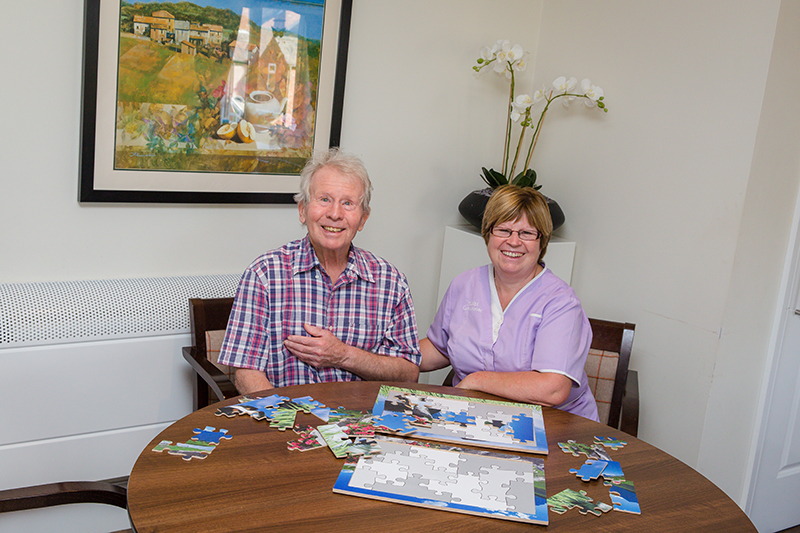 A male resident smiling with a female carer wearing a purple uniform playing with a puzzle. An plant in the background and a painting on the wall.