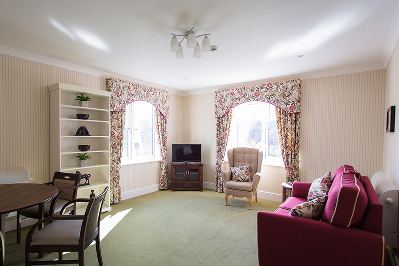Floral curtains with matching floral pillows. Red sofa and cream armchair. TV on stand. Bookcase with ornaments. Dining table. Green carpet and cream stripe wallpaper.