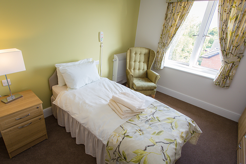 A light green feature wall. A Single bed with a floral bed runner with matching curtains. A green armchair in corner. Bedside table with a lamp turned on. Some towels on bed.
