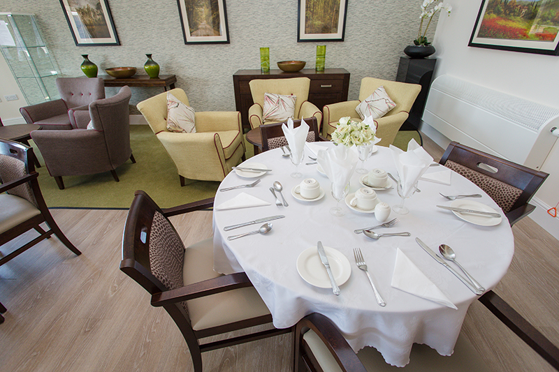 A dressed dining room table with cutlery and side plates out, as well as wine glasses and cups. Seating in the background with a coffee table.