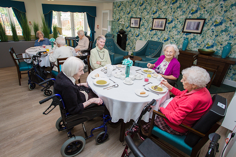 Two dressed tables. Smiling residents chatting and eating soup. A blue floral wallpaper in the background with three photoframes.