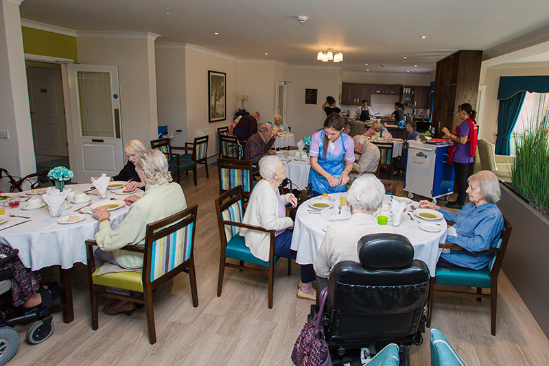 Five tables of residents eating soup for a starter. A female staff member speaking to a female resident. Another staff member in the background working. Chef in the corner serving food.