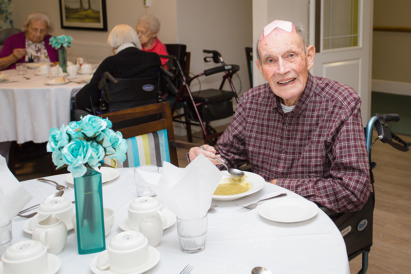 A smiling male resident in a wheel chair eating soup with blue flower on the dressed dining room table.