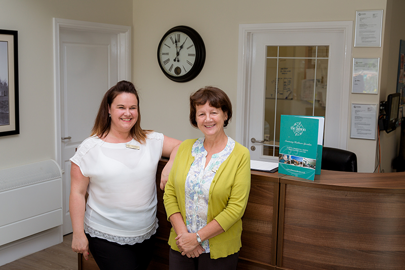 Two smiling female staff members in The Ashton Care Home reception area.