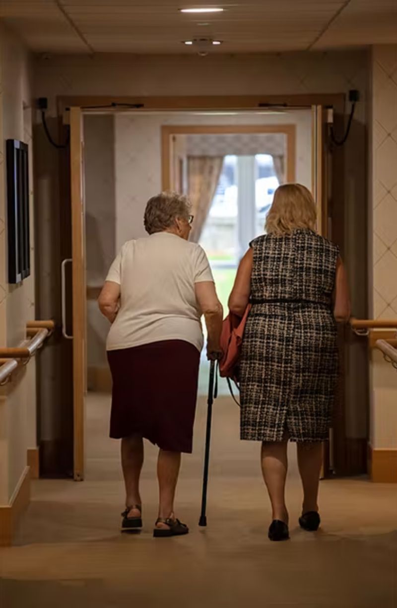 A female in a dress and a female resident with a cane walking down the hallway together.