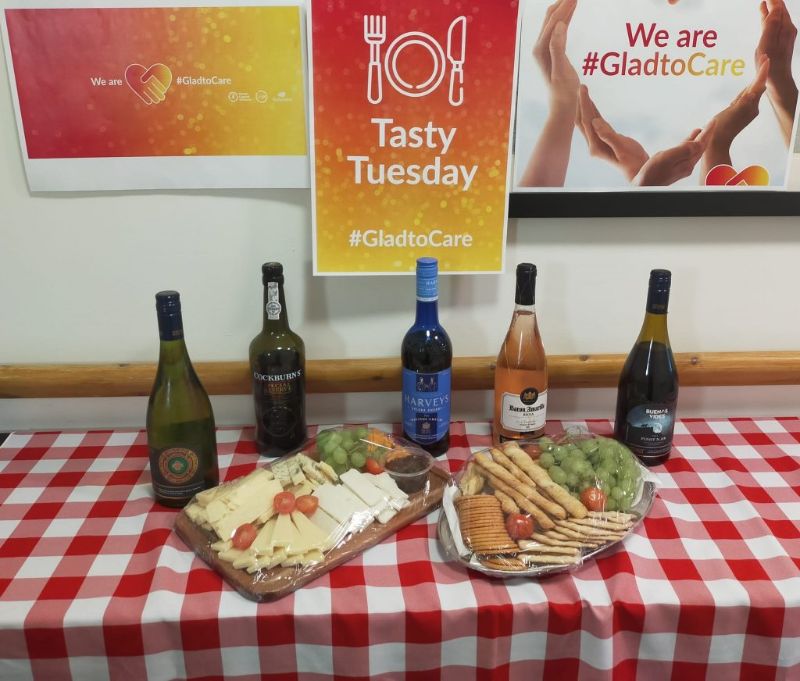 A red/yellow sign saying Tasty Tuesday with a selection of wines and two cheese boards on a red and white pattern table cover.