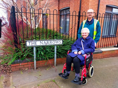 A female residents in a wheelchair with a male caregiver having a photo at a roadsign named "The Narrows".