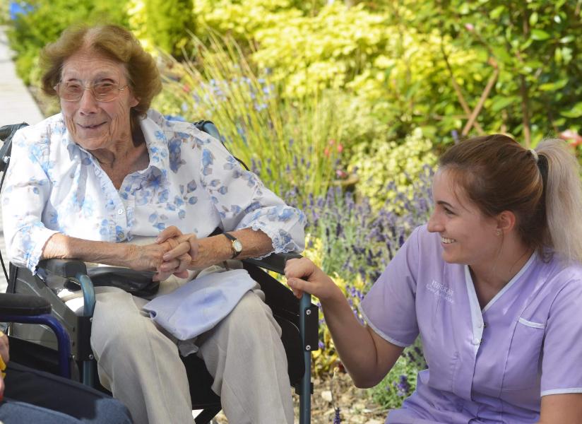 An elderly lady seated in a chair with a nurse kneeling beside her and smiling.