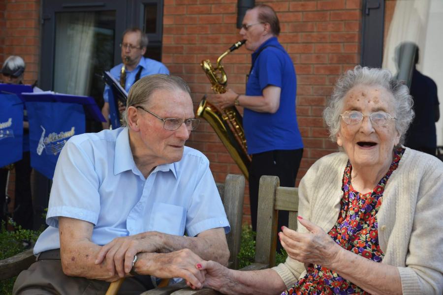 An elderly couple holding hands while a man plays the saxophone in the background.