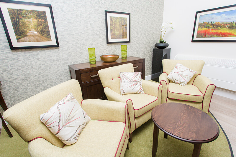 Three cream/yellow armchairs with floral pillows. Brown coffee table. Green rug. Pattern wallpaper and three painting on the wall.