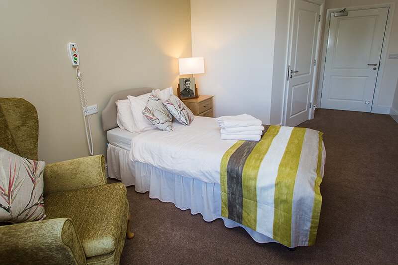 Brown carpet. Green feature wall. Bed with nurses remote. Green bed runner and floral pillows. Towel on bed. Bedside table with photo and lamp. Green armchair with floral pillow.