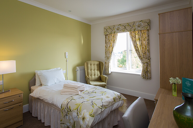 Green feature wall. Bed with floral bed runner and matching curtains. Green armchair in front of radiator. Bedside table with light turn on. Wardrobe and vanity with a vase and flowers.