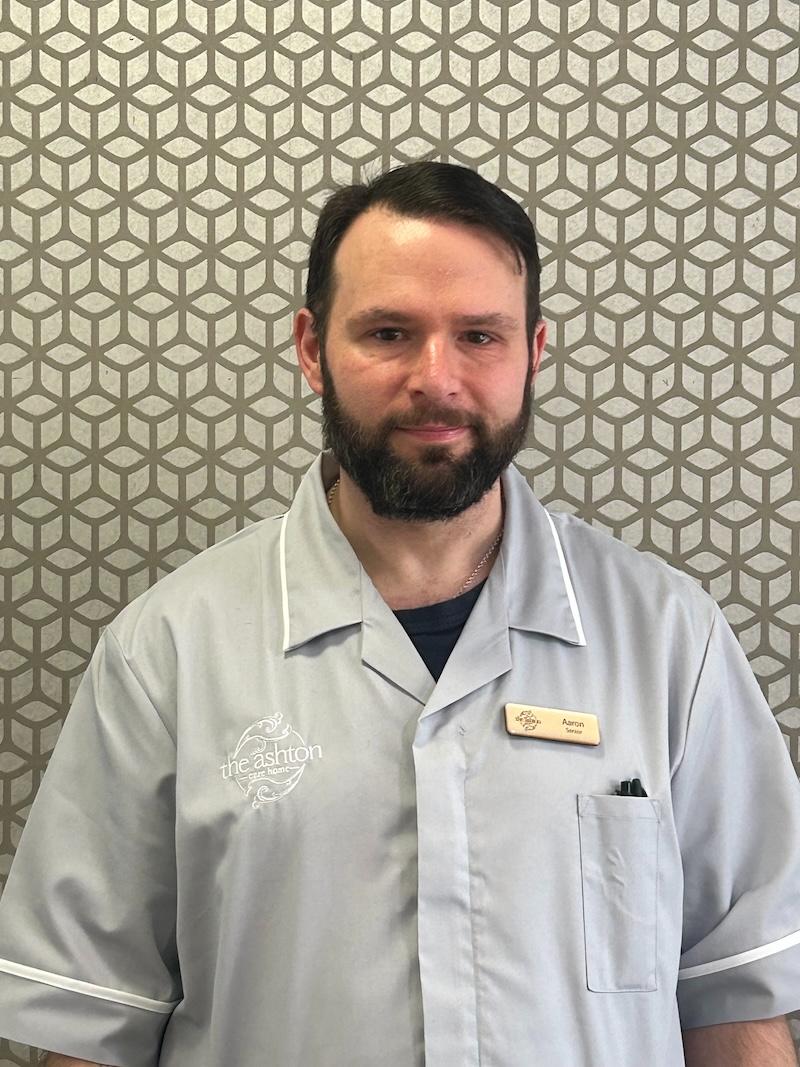 man with brown hair wearing a blue senior care assistant uniform having his photo taken for the team page