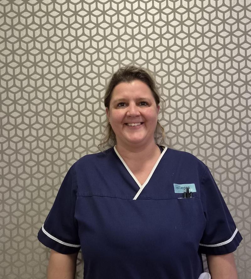 woman with brown hair wearing a navy nurses uniform stood smiling having her photo taken for the meet the team page
