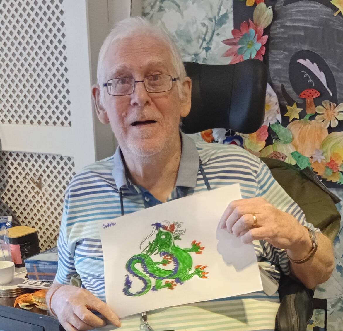 older man sat down holding up a drawing of a green chinese new year dragon