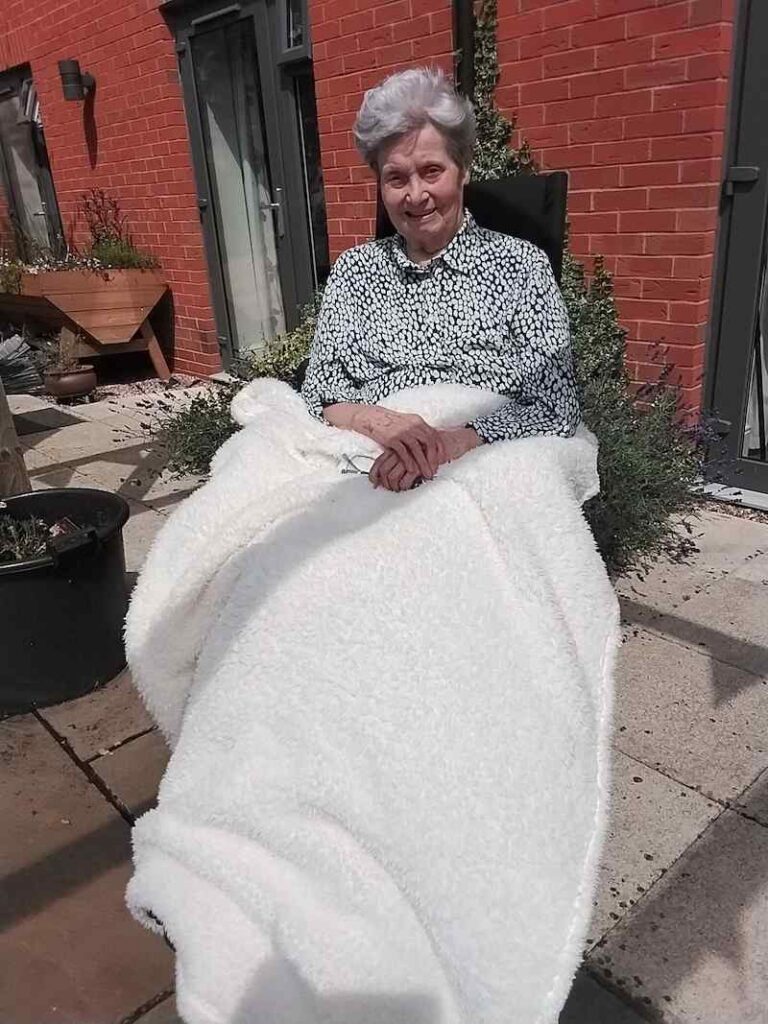 female resident sat in the garden with a white blanket on her lap smiling